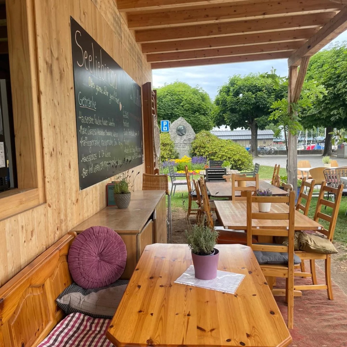Gedeckte Terrasse der Seeliebi mit Holzbank, Kissen und Lavendel auf dem Tisch, Tafel-Menü an der Wand und Blick in den Park am Bodensee